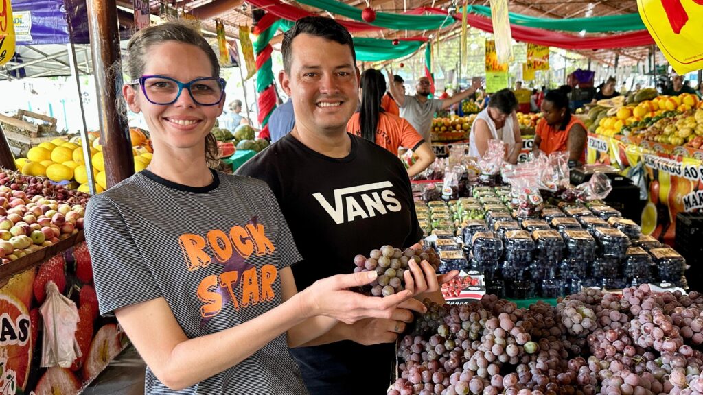 A imagem mostra um casal sorrindo em um mercado de frutas. A mulher, à esquerda, usa óculos azuis e uma camiseta listrada com a estampa "ROCK STAR". O homem, à direita, veste uma camiseta preta com o logo da Vans e segura um cacho de uvas. Eles estão em frente a uma barraca repleta de frutas frescas, com caixas de uvas embaladas e cachos de uvas maiores em exposição. Ao fundo, outras pessoas circulam pelo mercado, e há decorações coloridas com fitas verdes e vermelhas. A variedade de frutas, como melões, morangos e uvas, sugere um ambiente vibrante e abastecido.
