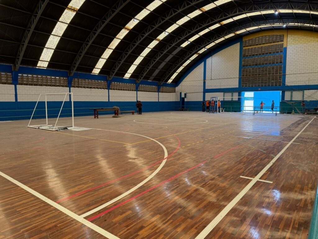 A imagem mostra um ginásio esportivo coberto, com piso de madeira polido marcado por linhas de diferentes modalidades, como basquete, vôlei e futsal. À esquerda, há uma trave branca posicionada, enquanto ao fundo algumas pessoas parecem inspecionar ou trabalhar no espaço. O ambiente tem teto alto em arco, com painéis de iluminação e paredes pintadas em azul e branco, transmitindo a ideia de um local multiuso preparado para treinos, jogos ou eventos esportivos.