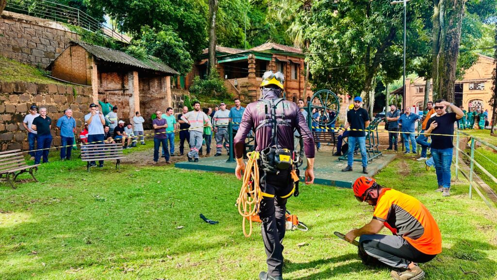 A imagem mostra um grupo de homens reunidos em um gramado, com algumas construções rústicas e árvores ao fundo. No primeiro plano, dois homens estão em destaque. Um deles, de costas para a câmera, veste um traje de escalada com cordas e equipamentos pendurados, e um capacete amarelo. Ele parece estar se preparando para alguma atividade. O outro homem, agachado, usa um capacete laranja e uma camiseta de manga curta laranja e cinza, e está manuseando uma motosserra. O restante do grupo, em pé e sentados em bancos, observa a cena. Eles parecem ser espectadores ou participantes de uma demonstração ou treinamento. As construções ao fundo sugerem um local histórico ou rural, com paredes de pedra e tijolo aparente, e telhados desgastados. A vegetação densa e as árvores compõem um cenário natural e sombreado. Há também uma estrutura metálica com uma grande roda no centro-direita da imagem. A atmosfera geral é de um evento ao ar livre, possivelmente relacionado a arborismo, segurança ou manutenção de áreas verdes.