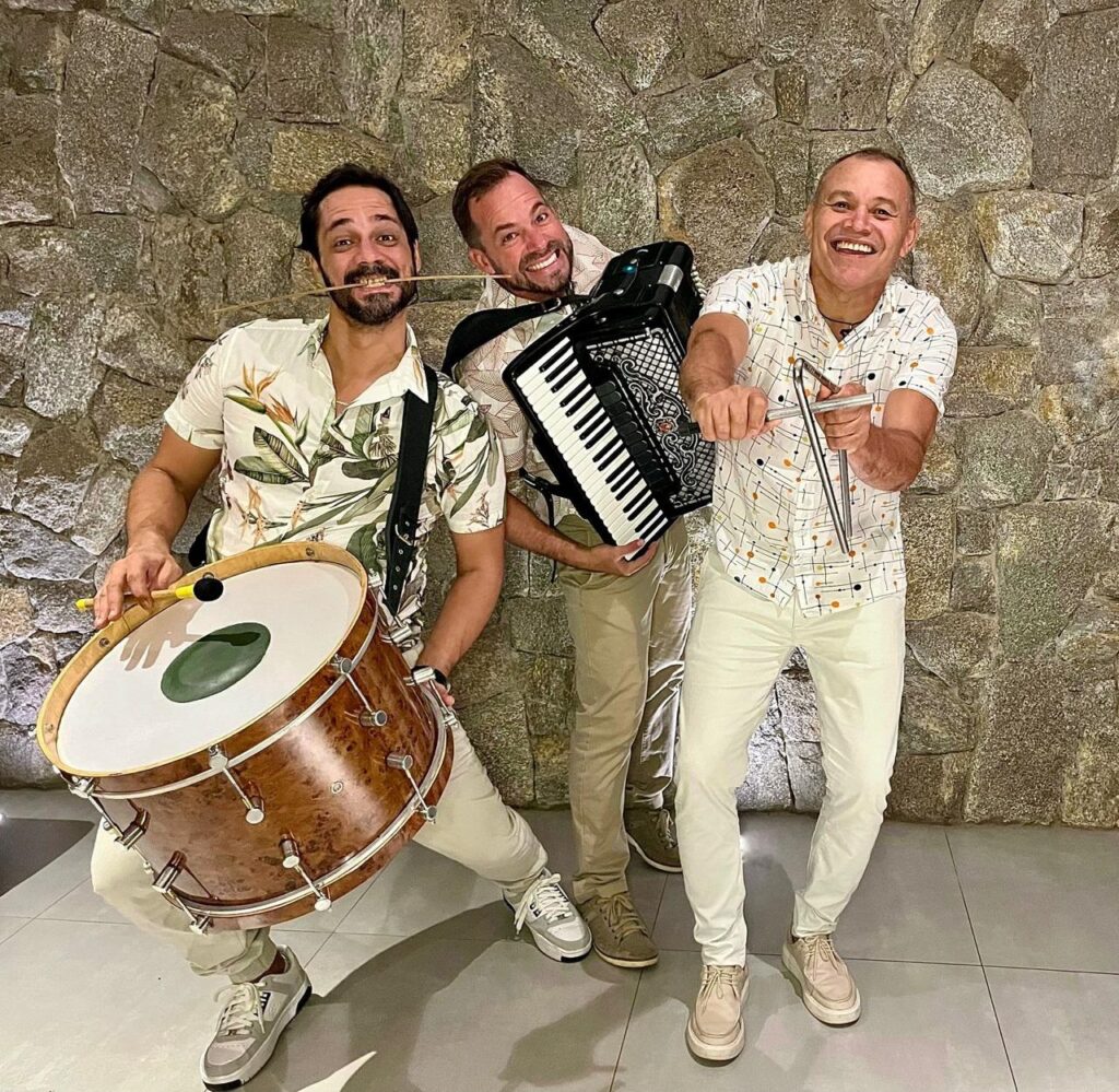 A imagem mostra três homens sorrindo e posando com instrumentos musicais em frente a uma parede de pedra. O homem da esquerda está usando uma camisa com estampa tropical e segura um tambor grande, com uma baqueta na boca. O homem do meio, vestindo uma camisa bege, toca um acordeão. O homem da direita, com uma camisa branca estampada, segura um instrumento de percussão feito de metal. Todos os homens estão vestidos com calças claras e tênis, e a atmosfera geral é de alegria e descontração.