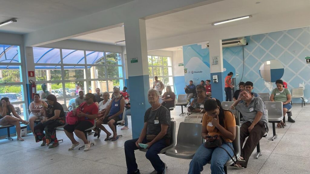 A imagem mostra uma sala de espera com várias pessoas sentadas em cadeiras, aguardando atendimento. A sala parece ser de uma unidade de saúde, com janelas grandes ao fundo que permitem a entrada de luz natural e mostram árvores e carros do lado de fora. As pessoas na sala de espera são de diversas idades e etnias, vestindo roupas casuais. Algumas estão concentradas em seus celulares, outras conversam entre si ou olham para frente. Há também alguns funcionários, como uma mulher de laranja que parece estar operando um dispensador de água ou lanche. A decoração da sala é simples, com paredes azuis e brancas e um padrão geométrico nas paredes. Há também um extintor de incêndio vermelho visível em um dos cantos. A iluminação da sala é feita por lâmpadas fluores…