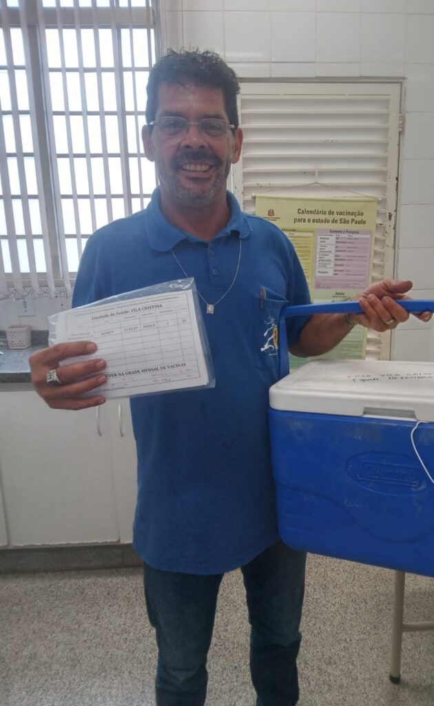 Um homem está segurando um cartão de registro de vacinação e uma caixa térmica azul. Ele está vestindo uma camiseta polo azul e jeans, além de óculos. O cartão de vacinação é da "Unidade de Saúde: VILA CRISTINA" e mostra detalhes sobre uma vacina da Pfizer administrada em "31.03.27". Ao fundo, há um cartaz intitulado "Calendário de vacinação para o estado de São Paulo". A caixa térmica parece ser da marca Coleman.
