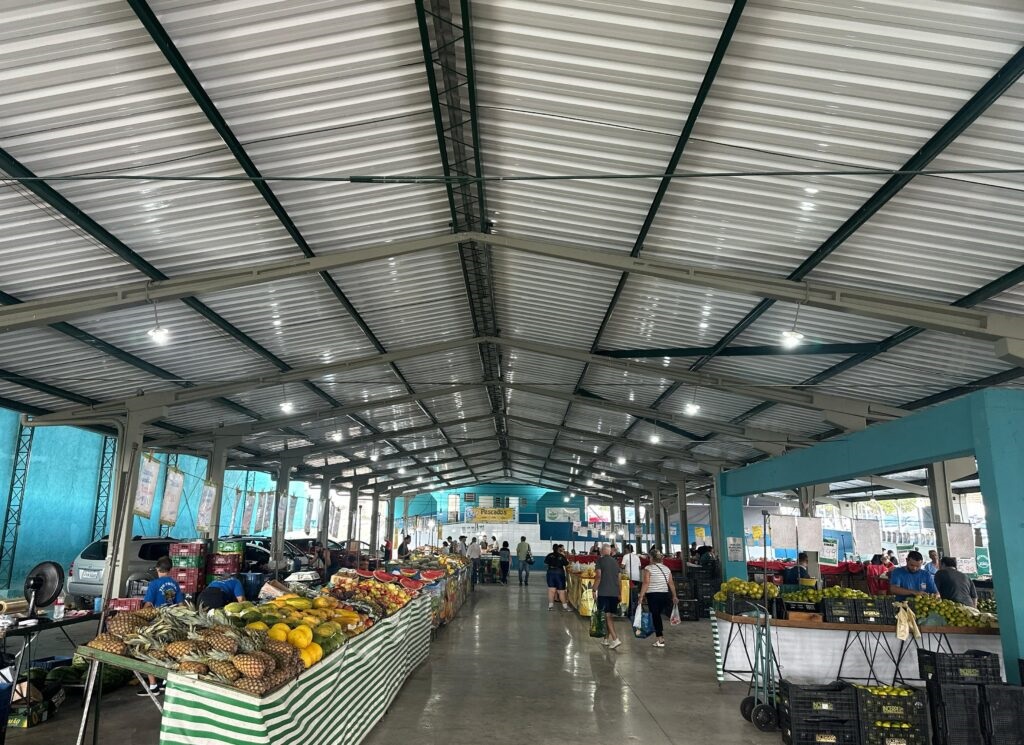 A imagem mostra um mercado ao ar livre com uma cobertura de telhado metálico. O mercado está repleto de barracas de frutas, com uma grande variedade de produtos expostos em mesas. As frutas mais visíveis incluem abacaxis, mangas, melancias e laranjas. Há pessoas circulando pelo mercado, algumas comprando e outras trabalhando nas barracas. A iluminação é feita por lâmpadas penduradas no teto, e a estrutura do mercado é composta por colunas e vigas metálicas. O chão é de cimento, e ao fundo, é possível ver carros estacionados e algumas construções.