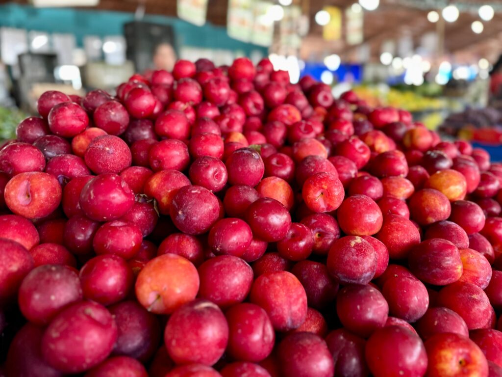 A imagem mostra uma grande quantidade de ameixas vermelhas e rosadas, empilhadas em uma feira ou mercado. As frutas estão em close-up, com foco nas que estão mais próximas da câmera, e o fundo está desfocado, mostrando o ambiente da feira com luzes e outras barracas. As ameixas apresentam diferentes tons de vermelho, com algumas tendo manchas alaranjadas, indicando que estão maduras e prontas para consumo. A textura da pele das frutas é visível, com algumas apresentando um leve brilho.