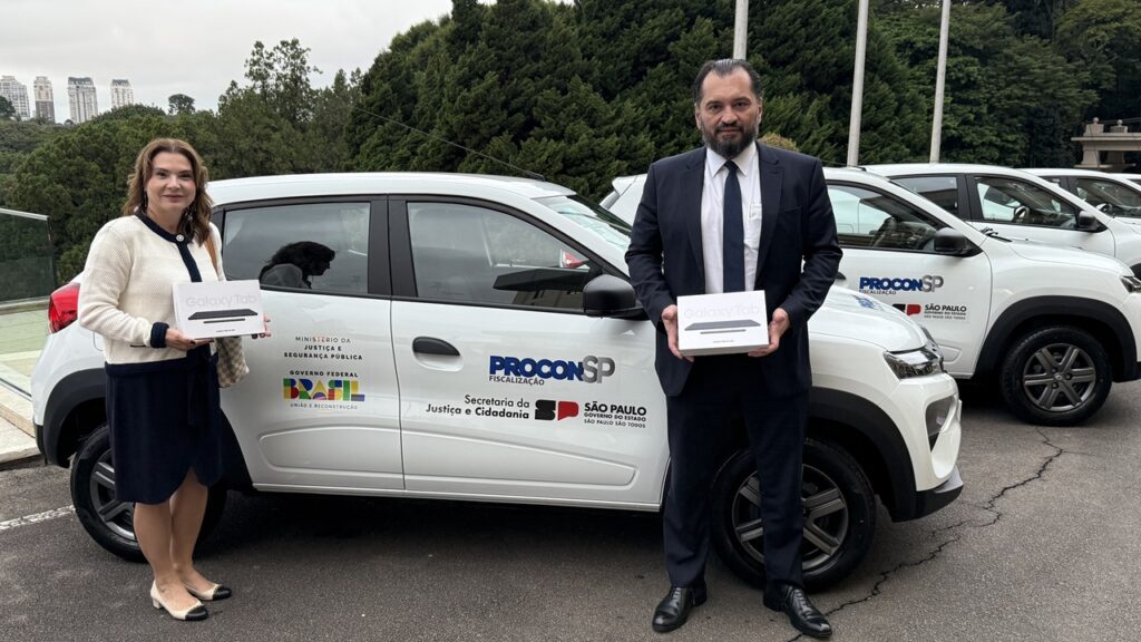 A foto mostra duas pessoas em frente a um veículo branco identificado com logotipos e inscrições de órgãos governamentais ligados à Justiça e ao Governo de São Paulo. Cada uma delas segura uma caixa contendo um tablet Samsung Galaxy Tab. Ao fundo, é possível ver outros veículos semelhantes alinhados, compondo uma frota. O cenário é externo, com árvores e prédios ao longe, e transmite a ideia de um evento oficial de entrega ou apresentação de equipamentos destinados a ações de fiscalização ou serviços públicos.