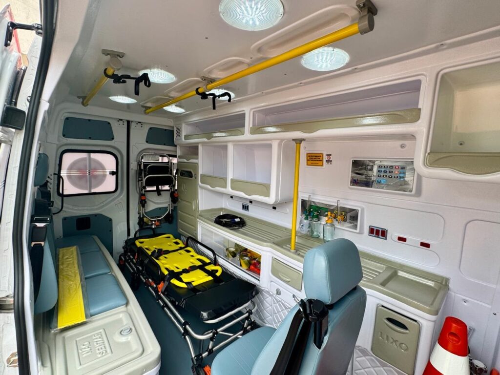 No lado esquerdo, há um banco azul claro com um encosto e um assento. Ao lado do banco, há um compartimento com a inscrição "NÃO SENTAR" (não sentar). No centro, destaca-se uma maca amarela com estrutura metálica, pronta para o transporte de pacientes. Acima da maca, há barras de apoio amarelas no teto, juntamente com luminárias circulares. À direita, observa-se um balcão com diversos equipamentos, incluindo frascos com líquidos, um painel de controle digital com botões e uma tela, e um pequeno lavatório. Há também um assento azul claro com encosto, posicionado mais à frente. Na parte superior, prateleiras abertas oferecem espaço de armazenamento. A iluminação geral do veículo é clara e uniforme. Na parte inferior direita, há um cesto de lixo com a inscrição "LIXO" e um cone de sinalização laranja e branco. O ambiente é limpo e organizado, projetado para o atendimento médico de emergência.
