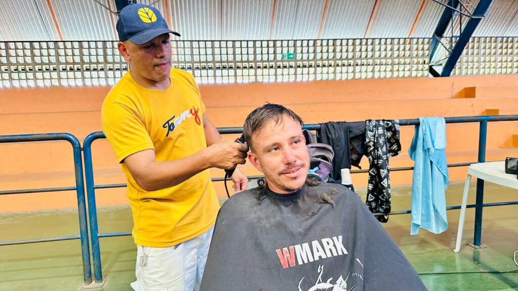 A imagem mostra dois homens em um ambiente interno, possivelmente uma quadra esportiva, com arquibancadas ao fundo. Um homem, vestindo uma camiseta amarela e um boné azul com um símbolo de folha, está cortando o cabelo de outro homem. Ele usa uma máquina de cortar cabelo elétrica, e o homem que está sendo cortado está sentado, usando uma capa preta com a inscrição "WMARK" em vermelho. O homem que está recebendo o corte de cabelo tem uma expressão levemente sorridente e parece estar olhando para o lado. Há alguns cabelos cortados espalhados sobre sua capa. Pendurados em uma grade metálica ao fundo, há algumas roupas, incluindo uma toalha azul e uma camiseta preta com um padrão de tesouras. Uma mesa branca com um objeto preto em cima está visível à direita. O chão parece ser de cor verde.