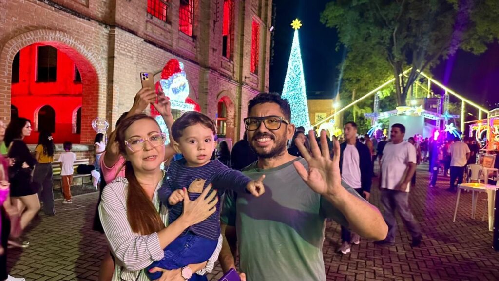 A família está em pé, em frente a um prédio de tijolos vermelhos, com janelas iluminadas em vermelho. Há decorações de Natal, incluindo uma árvore de Natal iluminada e uma figura de Papai Noel. A família parece estar se divertindo, e o homem acena para a câmera. Há outras pessoas no fundo.