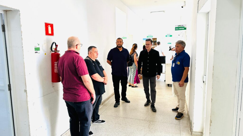A imagem mostra um grupo de pessoas em um corredor que parece ser de um hospital ou centro de atendimento. Há um extintor de incêndio vermelho na parede à esquerda, com um sinal vermelho acima dele. Quatro homens estão em primeiro plano, conversando. Um deles, vestindo uma camisa vinho, está de costas para a câmera. Ao lado dele, um homem com uma camisa preta e óculos. Em pé, atrás deles, um homem em uma camisa azul marinho escura. O quarto homem, à direita, veste uma camisa preta e calças cinzas. Mais atrás, no corredor, há outras pessoas e sinais verdes no alto, indicando "UPA 24h" e "CALL CENTER". Um homem com uma camisa azul está encostado na parede à direita, olhando para frente. O corredor é iluminado e o chão é de granilite.