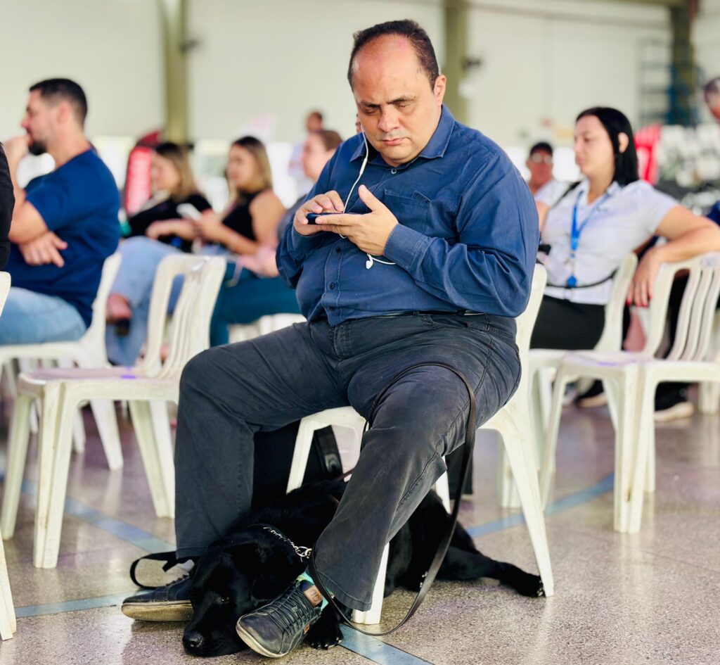 A imagem mostra um homem sentado em uma cadeira branca, usando fones de ouvido e olhando para seu celular. Ele está vestindo uma camisa azul e calças escuras. Abaixo da cadeira, um cachorro preto está deitado no chão, com a cabeça apoiada no pé do homem. Ao redor, outras pessoas estão sentadas em cadeiras, parecendo assistir a alguma apresentação ou evento. O ambiente parece ser um local fechado, possivelmente um auditório ou sala de eventos, com iluminação artificial.