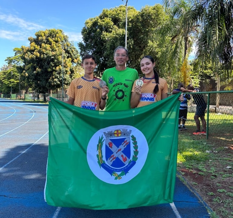 A imagem mostra três pessoas em pé, duas jovens e um homem mais velho, segurando uma bandeira verde com um brasão no centro. Os dois jovens, um rapaz e uma moça, usam medalhas no pescoço e seguram medalhas nas mãos. Ambos vestem camisetas com a inscrição "WORLD TENNIS". O homem mais velho, atrás deles, veste uma camiseta verde com um círculo e uma inscrição no peito. A bandeira é grande e ocupa a parte inferior da imagem. O brasão no centro é circular e contém um escudo com símbolos azuis e vermelhos, cercado por ramos verdes e uma inscrição em latim. Ao fundo, observa-se uma pista de atletismo azul, árvores frondosas e um céu azul claro. Há também outras pessoas ao fundo, perto de uma grade. A cena parece ser um evento esportivo ao ar livre, possivelmente uma competição de tênis, onde os jovens foram premiados.
