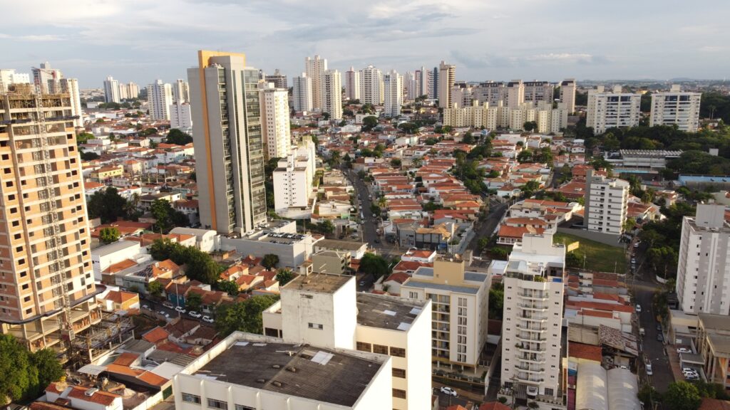 Imagem aérea de Piracicaba: de um lado, imponentes edifícios de vidro e múltiplos andares refletem o dinamismo urbano; de outro, casas baixas com telhados vermelhos, dispostas em linhas organizadas, preservam um ar acolhedor e residencial. As ruas arborizadas, repletas de veículos, conectam esses dois mundos, revelando uma convivência harmoniosa entre a verticalidade da arquitetura contemporânea e a horizontalidade dos bairros tradicionais, compondo um cenário vibrante de diversidade arquitetônica e desenvolvimento urbano.