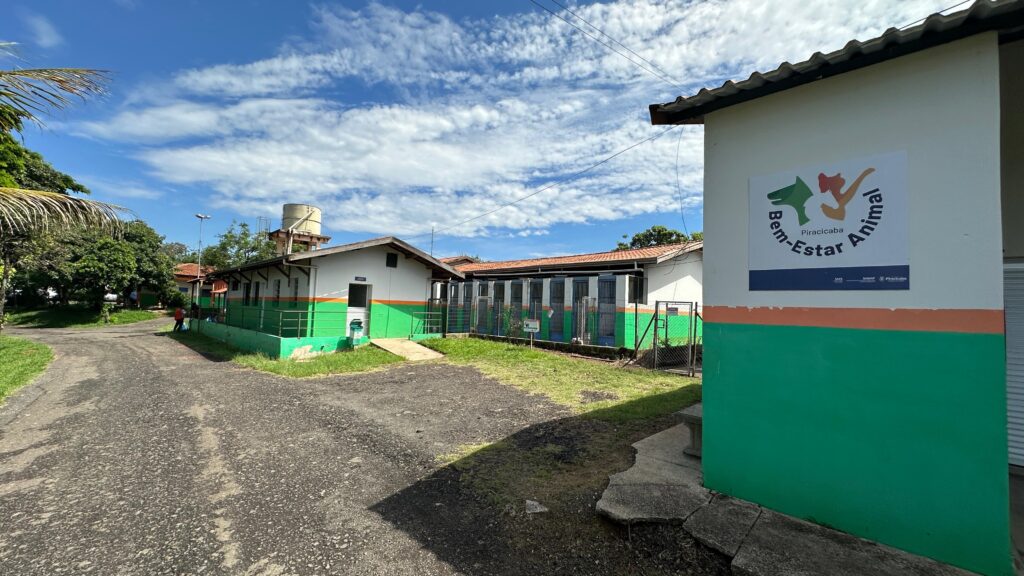 A imagem mostra um centro de bem-estar animal com um céu azul claro e nuvens brancas ao fundo. O edifício principal é branco com detalhes em verde e laranja. Há uma placa na parede com o logotipo "Bem-Estar Animal Piracicaba" e os logotipos do SMS, SIMAP e Piracicaba. À esquerda do edifício principal, há um caminho de asfalto que leva a uma área gramada com árvores. Há também uma torre de água visível ao fundo. O edifício parece ser um abrigo para animais, com várias baias visíveis ao longo do lado direito da estrutura. A vegetação exuberante e o céu claro sugerem um dia ensolarado.