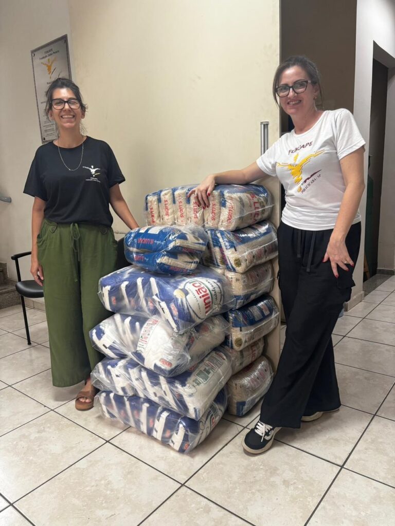 A imagem mostra duas mulheres em pé ao lado de uma pilha de pacotes de arroz. A mulher à esquerda veste uma camiseta preta com um logotipo branco e calças verdes folgadas. Ela usa óculos e tem um sorriso largo. A mulher à direita veste uma camiseta branca com um logotipo amarelo e preto e calças pretas. Ela também usa óculos e tem um sorriso. A pilha de arroz é alta e parece conter muitos pacotes. Os pacotes têm embalagens brancas e azuis com texto em português.