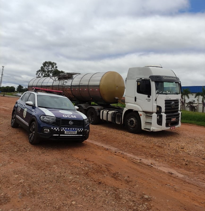A imagem mostra um veículo oficial da Guarda Civil Municipal (GCM) estacionado à frente de um caminhão-tanque. O carro da GCM é um SUV azul escuro com detalhes em branco e azul claro, e possui um giroflex vermelho no teto. A palavra "GCM" está escrita em destaque nas laterais e na frente do veículo, juntamente com o brasão da instituição. A placa do carro é "SUM0G90". O caminhão-tanque é branco e de grande porte, com o tanque prateado e uma seção amarelada na parte traseira. Há uma inscrição "Constellation" no topo da cabine e "Fontana" escrita na lateral da cabine. O caminhão está estacionado em uma estrada de terra com vegetação ao fundo e um céu nublado. A cena sugere uma fiscalização ou parada de rotina envolvendo o veículo oficial e o transporte de carga.