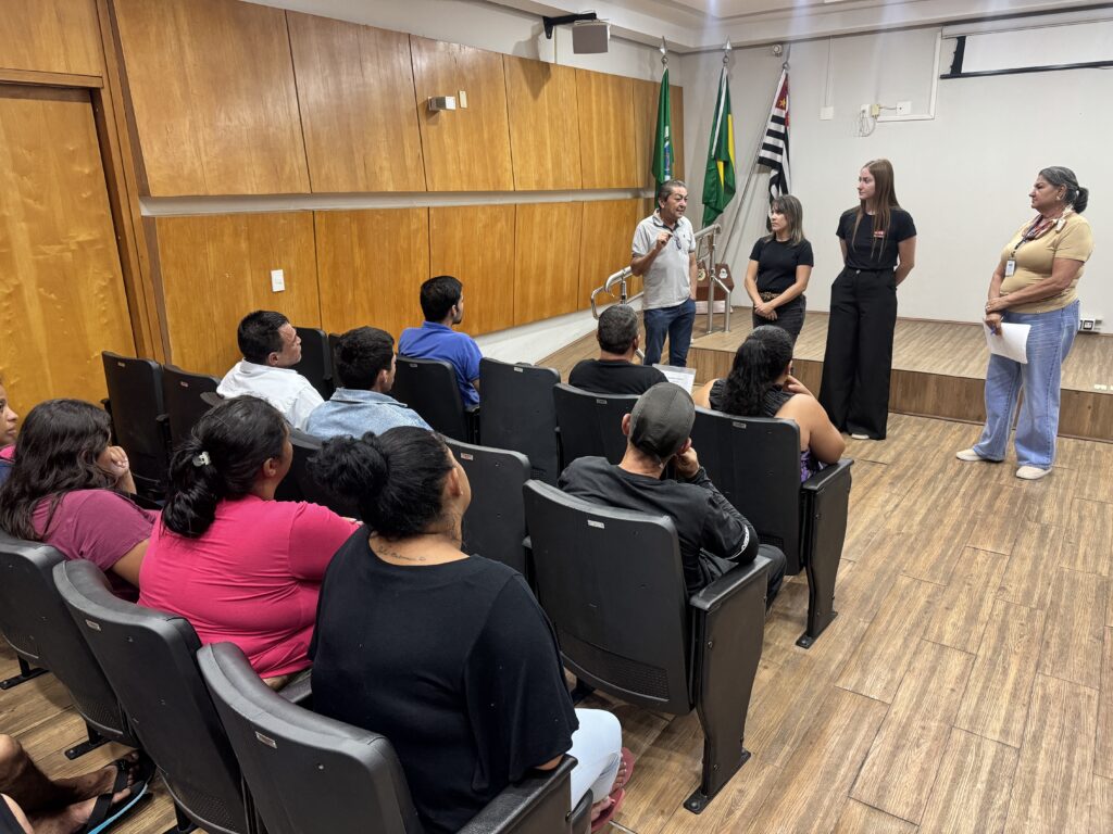 A imagem mostra uma sala de reunião ou auditório, com um grupo de pessoas sentadas em cadeiras voltadas para um palco ou área elevada. No palco, há quatro pessoas em pé. Um homem, vestindo uma camisa cinza e calça jeans, está de pé no centro, gesticulando enquanto fala. À sua direita, há uma mulher com uma camisa preta e calça xadrez. Mais à direita, outra mulher, com uma camisa preta e calça preta, está de pé. A quarta pessoa, uma mulher com uma camisa bege e calça jeans, está mais afastada, segurando um papel. A sala possui paredes revestidas de madeira, com um design em painéis. Há três bandeiras hasteadas atrás do palco, com as cores verde, amarela, preta e branca. O chão é de madeira clara, com um padrão de tábuas. As cadeiras onde o público está sentado são pretas, e algumas pessoas estão olhando para o palco, enquanto outras parecem conversar ou descansar. A iluminação da sala parece ser artificial, vindo de luminárias no teto.