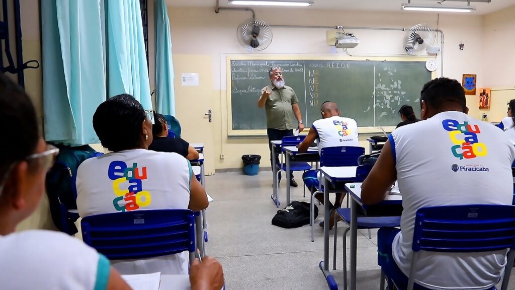 A imagem mostra uma sala de aula com um professor explicando algo no quadro negro para os alunos. Os alunos estão sentados em carteiras, voltados para o quadro. Alguns alunos estão usando camisetas brancas com um logo colorido escrito "Educação" e "Piracicaba". O professor, um homem com barba, está de pé em frente ao quadro e gesticula com a mão. No quadro, há algumas anotações em português, incluindo a data "28 de dezembro de 2023" e a palavra "VOCAIS: A, E, I, O, U", seguida por sílabas com as consoantes N e as vogais. A sala de aula parece ser simples, com cortinas azuis em uma parede e um ventilador de teto. Há também uma lixeira e alguns materiais escolares espalhados.