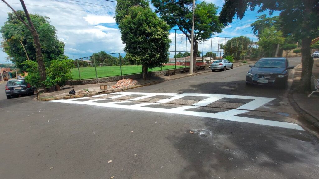 A imagem mostra uma cena urbana durante o dia, com um cruzamento destacado por faixas de pedestre recém-pintadas em branco. Em primeiro plano, há um carro hatchback escuro estacionado à esquerda, próximo a uma calçada onde há algumas pilhas de entulho de tijolos e caixas de papelão, indicando que pode ter ocorrido uma obra ou limpeza recente. Ao fundo, do lado esquerdo, é possível ver um campo de futebol com grama verde e traves, cercado por uma cerca. Mais adiante, na rua, há outros carros estacionados, incluindo um sedã prata e outro carro escuro do lado direito, próximo a uma árvore grande que projeta sombra sobre a via. Ao lado da árvore, um banco de praça está visível. O céu está parcialmente nublado, com nuvens brancas sobre um fundo azul, e a iluminação sugere que é começo ou meio de tarde. A cena é tranquila e parece ser um bairro residencial, com casas ao fundo e muita vegetação ao redor. O foco da imagem é o cruzamento, que conecta a calçada ao restante da rua, dando destaque à segurança dos pedestres.