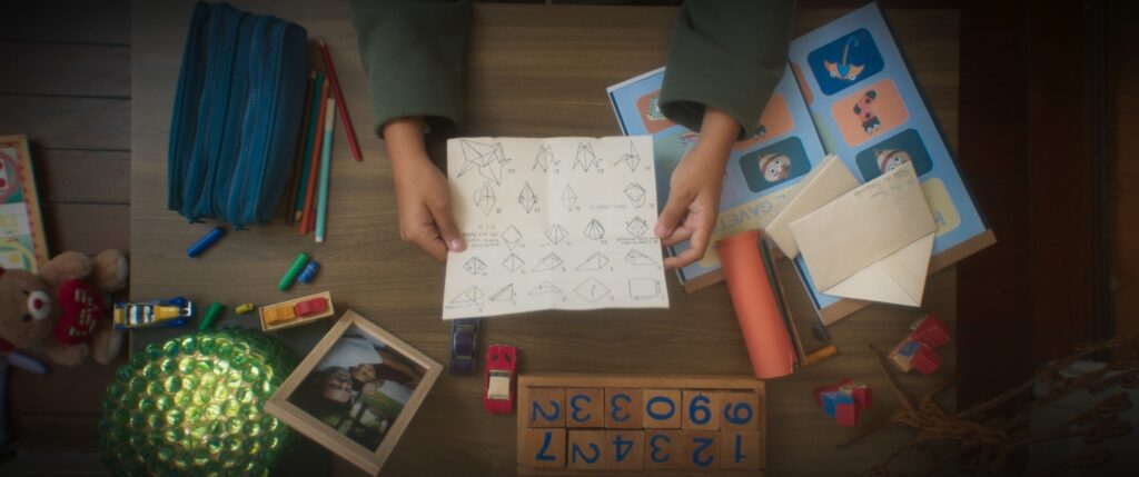 A imagem mostra uma mesa de madeira com vários objetos espalhados, como se fosse um cantinho de atividades de uma criança. No centro, uma criança segura um papel com instruções desenhadas para fazer origami. Ao redor, há materiais de arte como lápis de cor, um estojo azul, e um rolo de papel laranja. Há também brinquedos como carrinhos, um urso de pelúcia com um coração, blocos de montar e um conjunto de números em cubos de madeira. Uma moldura com uma foto de um casal e uma bola verde decorativa completam a cena, sugerindo um ambiente lúdico e criativo.