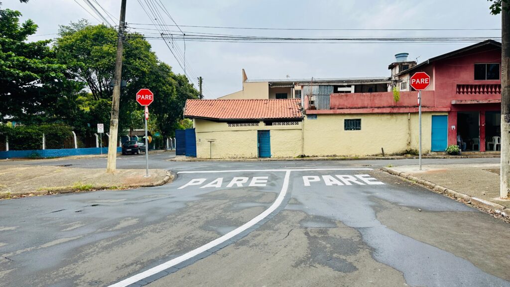 A imagem retrata um cruzamento de ruas em um dia nublado. No asfalto, a palavra "PARE" está pintada em letras brancas em ambas as direções de tráfego, indicando a necessidade de parada obrigatória. Sinais de "PARE" vermelhos, com a mesma palavra em branco, estão posicionados em postes em cada lado da rua, reforçando a sinalização horizontal. À esquerda, uma cerca azul delimita um terreno com vegetação densa e árvores altas. Um carro escuro está estacionado próximo a um poste de luz. À direita, edifícios de cores variadas compõem a paisagem urbana. Um prédio em tom de rosa vibrante se destaca, com janelas e uma varanda no andar superior. Ao lado, um edifício mais baixo de cor amarela apresenta um telhado de telhas vermelhas e uma porta azul. A atmosfera geral é de uma rua residencial ou comercial tranquila, com a sinalização indicando um ponto de atenção para os motoristas.
