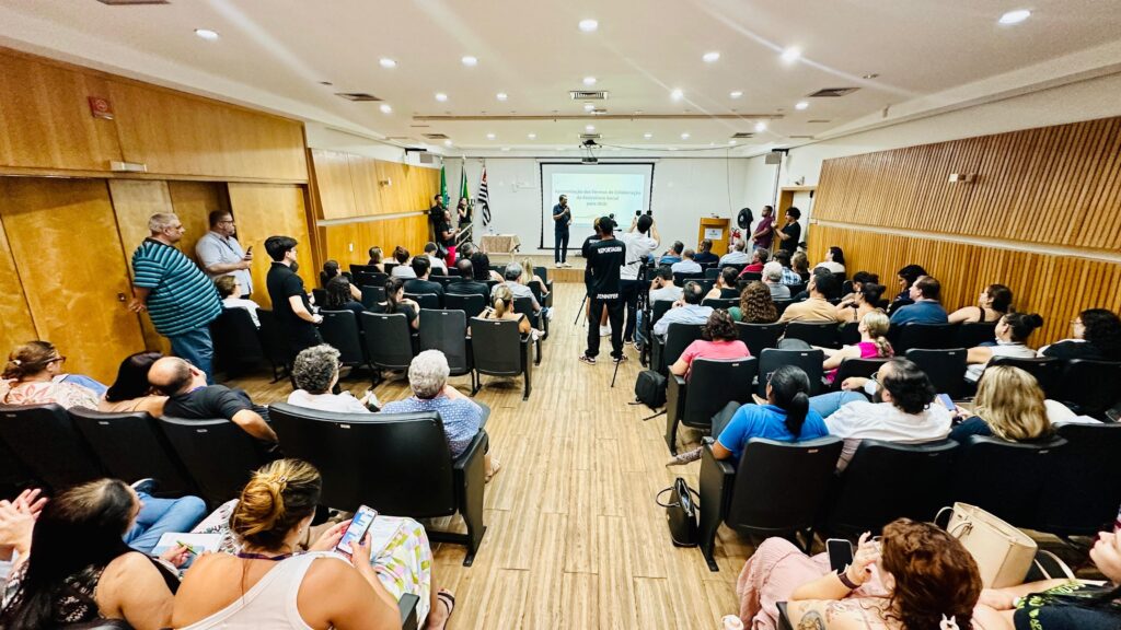 A imagem mostra um auditório com uma plateia sentada em cadeiras, voltada para um palco onde há um projetor exibindo um slide. No palco, algumas pessoas estão em pé, incluindo um homem que parece estar apresentando. Há bandeiras ao lado do palco. A iluminação é artificial, vinda de luminárias no teto. As paredes do auditório são revestidas de madeira clara e as cadeiras da plateia são pretas. Algumas pessoas na plateia estão com celulares ou papéis nas mãos.