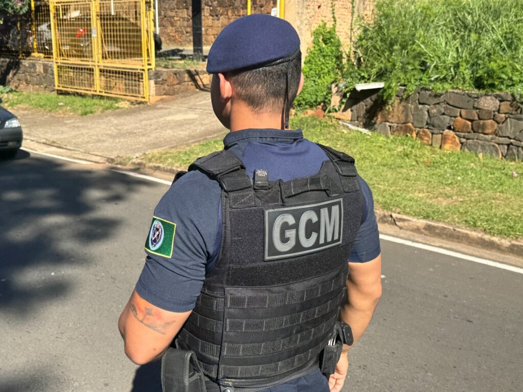 A imagem mostra um guarda municipal em serviço, visto de costas, em um dia ensolarado. Ele veste um uniforme azul escuro com um colete tático preto por cima. No colete, em destaque, está a sigla "GCM" em letras grandes e prateadas. Na manga esquerda do uniforme, há um distintivo circular com detalhes em verde e um símbolo no centro. O guarda usa um boné azul escuro e é possível notar uma tatuagem em seu braço direito. Ao fundo, vê-se uma rua asfaltada, um gramado, uma mureta de pedras e uma edificação com grades amarelas. A luz do sol cria sombras na rua, indicando que a foto foi tirada durante o dia.