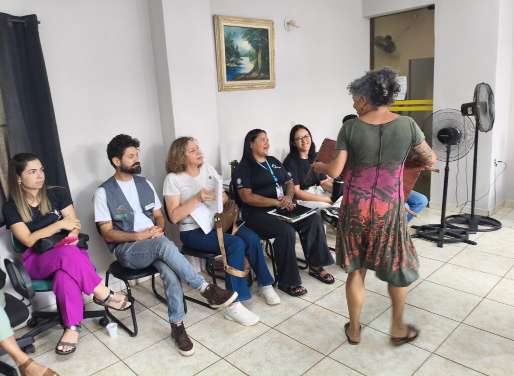 A imagem mostra um grupo de pessoas sentadas em cadeiras em uma sala, com uma mulher em pé de costas para a câmera, falando com o grupo. Ela está vestindo um vestido verde e rosa com um padrão floral. O grupo parece estar participando de uma reunião ou workshop. Há dois ventiladores em pé ao fundo, e uma pintura de paisagem emoldurada na parede. As pessoas sentadas estão vestindo roupas casuais, e algumas delas parecem estar segurando papéis ou cadernos. A iluminação na sala é clara, e o piso é revestido de azulejos.
