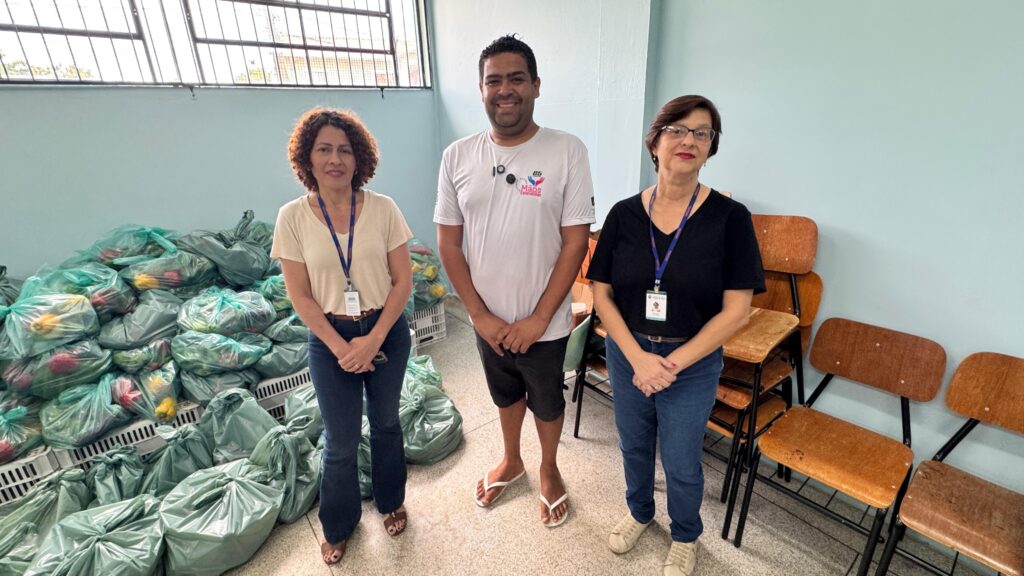 A foto mostra três pessoas em um ambiente interno, possivelmente uma sala comunitária ou escola. No centro, há um homem de camiseta branca com o logo "Mãos Estendidas" e shorts escuros. Ao lado dele, à esquerda, está uma mulher com cabelo cacheado, camisa clara e jeans, usando um crachá no pescoço. À direita do homem, há outra mulher com cabelo curto e óculos, vestindo camiseta preta, jeans e também com crachá. Todos estão sorrindo e olhando para a câmera. À esquerda da imagem, há várias sacolas verdes cheias de alimentos, como pimentões, sugerindo uma ação de doação ou distribuição de mantimentos. À direita, vê-se algumas cadeiras empilhadas encostadas na parede azul clara. A iluminação é clara, indicando que a foto foi tirada durante o dia. O ambiente transmite uma sensação de solidariedade e trabalho comunitário.