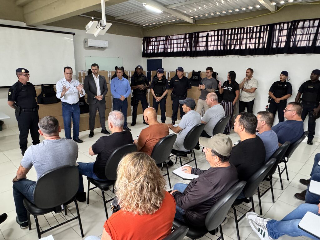 A imagem mostra uma reunião formal realizada em uma sala ampla com paredes brancas e teto de metal com iluminação fluorescente. À frente, há um grupo de pessoas, incluindo um homem de camisa azul clara que está falando, gesticulando com as mãos levantadas, e ao seu lado outros homens, alguns em uniformes táticos da GCM (Guarda Civil Municipal), usando boinas e coletes. Atrás deles, há várias caixas de papelão empilhadas e coletes táticos pretos sobre uma mesa. À frente, sentados em cadeiras pretas, estão cerca de uma dúzia de pessoas, a maioria homens, que parecem ser o público ou participantes da reunião. Eles estão atentos ao que está sendo apresentado, e alguns parecem estar tomando notas. O ambiente sugere uma apresentação ou entrega oficial de equipamentos para a GCM. A atmosfera é formal e profissional, com foco em segurança pública ou treinamento.