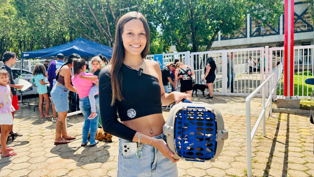A mulher sorri para a câmera enquanto segura um transportador de animais azul. Ela está usando um top preto de um ombro só, jeans e joias. atrás dela, há uma tenda azul, pessoas e uma cerca branca.