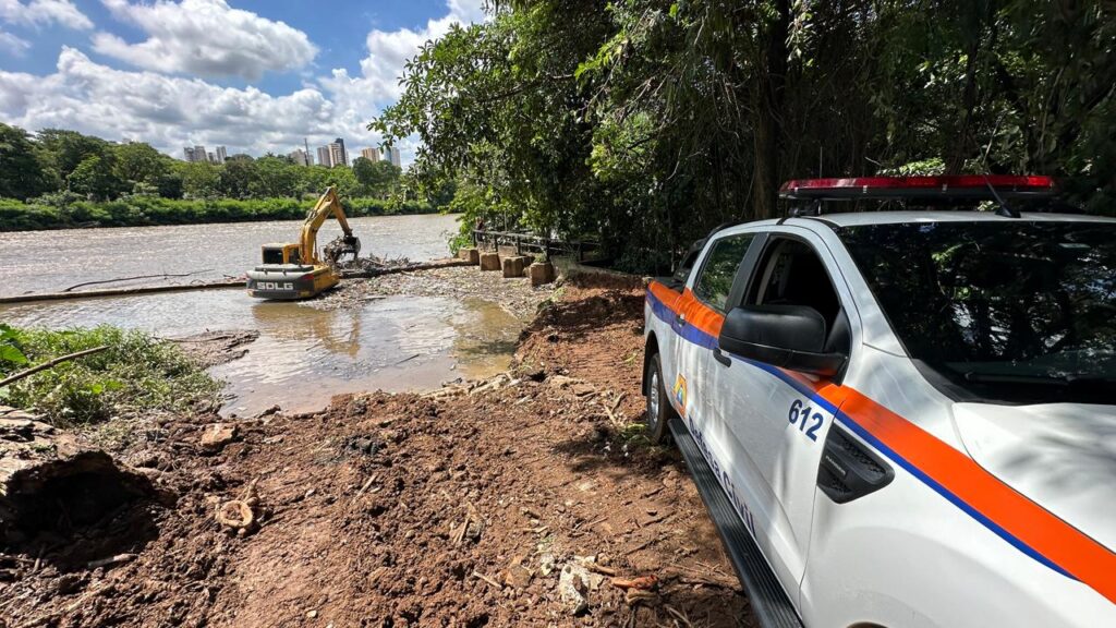 A cena mostra uma operação de defesa civil em um rio, onde um escavador amarelo da marca SDLG trabalha na água rasa, aparentemente removendo sedimentos ou detritos. À margem, um veículo branco identificado como “Defesa Civil” com sirene vermelha está estacionado sobre o terreno lamacento, reforçando o caráter emergencial da ação. Ao fundo, a paisagem combina áreas verdes com prédios altos sob um céu parcialmente nublado, sugerindo um esforço coordenado entre máquinas e equipes para prevenção de enchentes, limpeza ambiental ou manutenção da infraestrutura urbana.