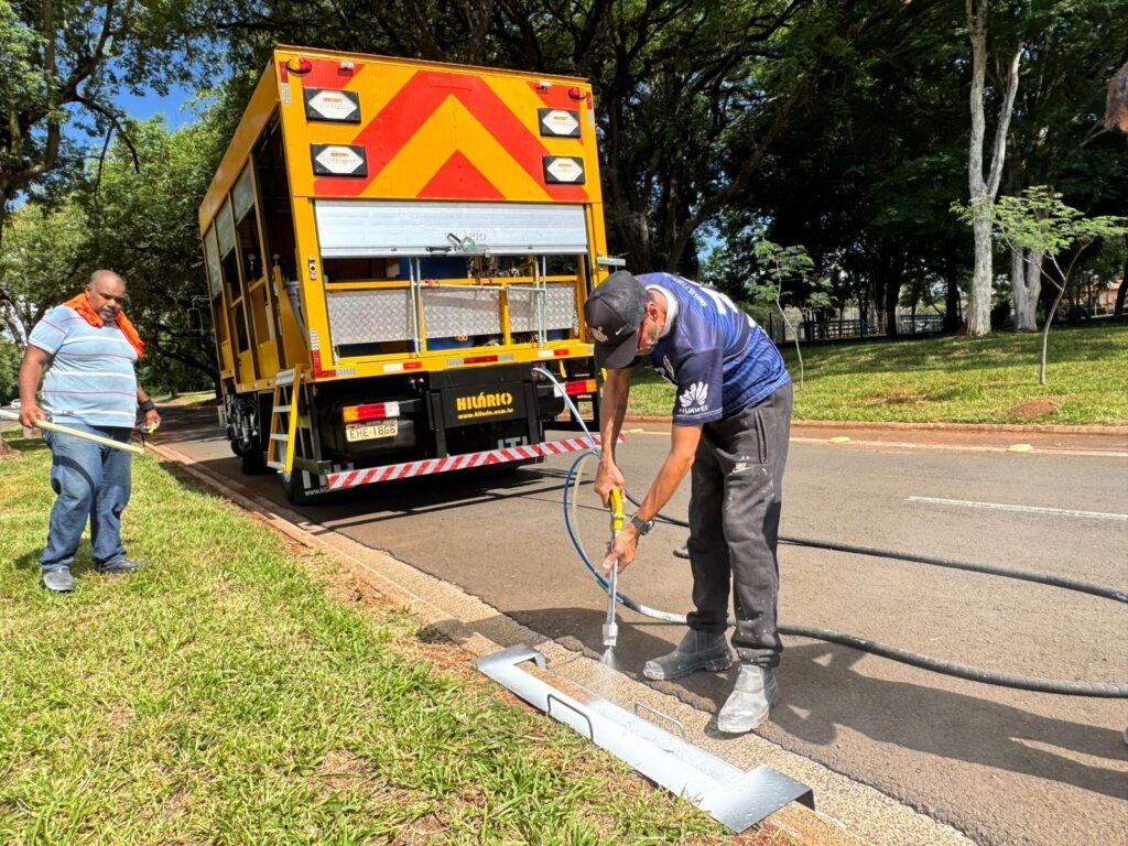 A imagem mostra dois homens em frente a um caminhão amarelo com detalhes em laranja e vermelho. O caminhão parece ser um veículo de serviço, possivelmente para manutenção de estradas ou sinalização. O homem à esquerda, vestindo uma camisa listrada azul e branca e jeans, segura um cabo de madeira. Ele está em pé na grama, perto da beira de uma estrada. O homem à direita, usando um boné preto, uma camiseta azul com um logo da Huawei e calças de moletom pretas, está ajoelhado na beira da estrada. Ele está operando uma ferramenta que parece ser um pulverizador ou um aplicador de algum tipo, direcionando-o para uma peça de metal prateada que está no chão. A ferramenta está conectada a uma mangueira preta que se estende até o caminhão. O fundo da imagem mostra árvores altas e exuberantes, sugerindo um ambiente externo, possivelmente um parque ou uma área arborizada ao longo de uma estrada. O céu está claro e azul. O caminhão tem a marca "HILÁRIO" e um site, "www.hilario.com.br". A placa do caminhão exibe "EHE-1865".