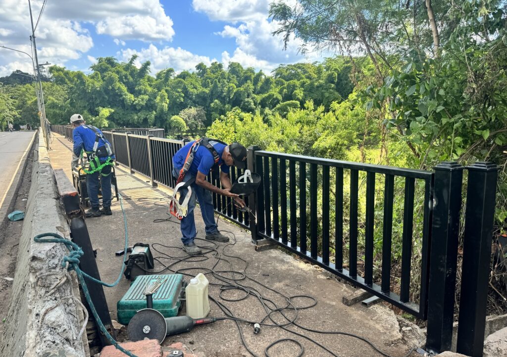 A imagem mostra dois trabalhadores em uma ponte, realizando reparos ou instalações em uma grade de proteção. Ambos os trabalhadores estão usando equipamentos de segurança, incluindo cintos de segurança e capacetes. O trabalhador mais próximo à câmera está ajoelhado e usando uma máscara de solda, indicando que ele está soldando a grade. Equipamentos de solda, como uma máquina de solda, cabos e um esmerilhador, estão espalhados no chão perto dele. Uma garrafa de líquido e uma caixa de ferramentas verde também estão visíveis. O outro trabalhador está em pé, mais ao fundo, e parece estar operando outra máquina, possivelmente um gerador ou outro equipamento elétrico. Ele também está usando um cinto de segurança e um capacete. A ponte parece estar ladeada por uma estrada em um lado e uma área verde exuberante com árvores altas do outro. O céu está parcialmente nublado, sugerindo um dia ensolarado. A grade de proteção é preta e parece ser feita de metal. O trabalho parece estar em andamento, com a grade ainda sendo instalada ou reparada.