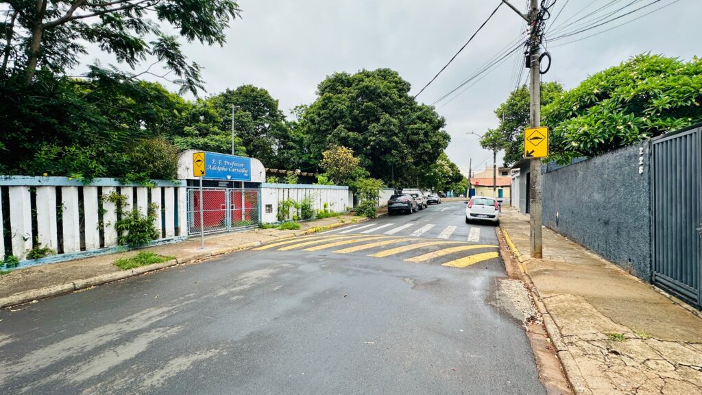 A imagem mostra uma rua em um dia nublado, com uma escola à esquerda e uma casa à direita. A rua tem uma faixa de pedestres com amarelos e amarelos listras, e um redutor de velocidade. Há carros estacionados na rua e na calçada. A escola tem um muro branco com azul e uma placa que diz "E. E. Professor Adolpho Carvalho". A casa tem um muro cinza e um portão de metal. Há árvores e vegetação em ambos os lados da rua.