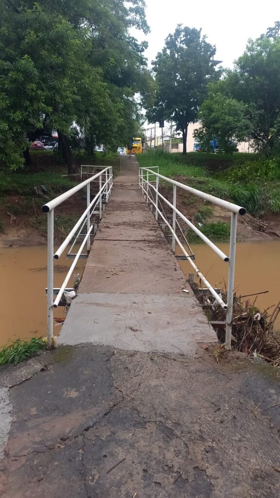 A imagem mostra uma ponte estreita para pedestres que atravessa um rio de água turva e marrom. A ponte é feita de concreto e possui corrimões metálicos brancos dos dois lados, embora estejam sujos e desgastados. O piso da ponte está coberto por lama seca e detritos, indicando que provavelmente houve uma enchente recente. No rio, é possível ver galhos e lixo presos nas estruturas da ponte. Ao fundo, há um caminhão amarelo e alguns carros estacionados ou em movimento, sugerindo que a ponte está situada próxima a uma área urbana ou suburbana. À esquerda, há árvores densas e verdes, enquanto à direita há um banco de terra com mais vegetação. A iluminação está opaca, com céu nublado, conferindo um aspecto cinzento e melancólico à cena. No centro da ponte, uma pessoa caminha para longe, quase desaparecendo na distância. A imagem transmite uma sensação de abandono e passagem do tempo, possivelmente após um evento climático intenso.