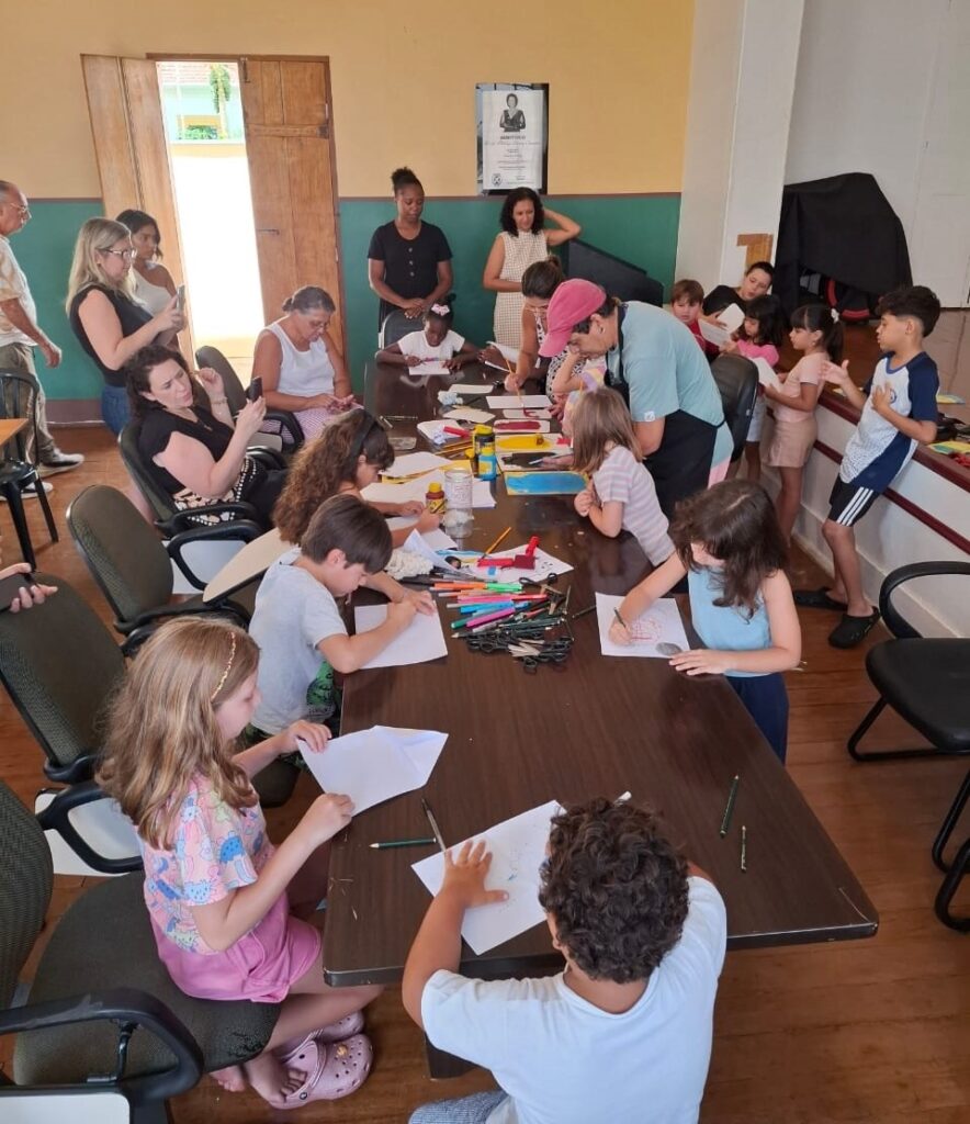 A imagem mostra um grupo de pessoas, incluindo adultos e crianças, reunidas em torno de uma mesa grande, aparentemente participando de uma atividade artística ou educativa. Há papéis, lápis de cor, canetas e outros materiais de arte espalhados pela mesa, sugerindo que estão desenhando ou pintando. Várias crianças estão sentadas à mesa, concentradas em seus trabalhos. Algumas crianças mais velhas e adultos estão em pé ou sentados nas cadeiras ao redor da mesa, observando ou participando da atividade. O ambiente parece ser um salão ou sala de aula, com paredes pintadas e uma porta aberta ao fundo. Há também um quadro de avisos na parede com uma foto emoldurada. A atmosfera geral parece ser de aprendizado e criatividade.