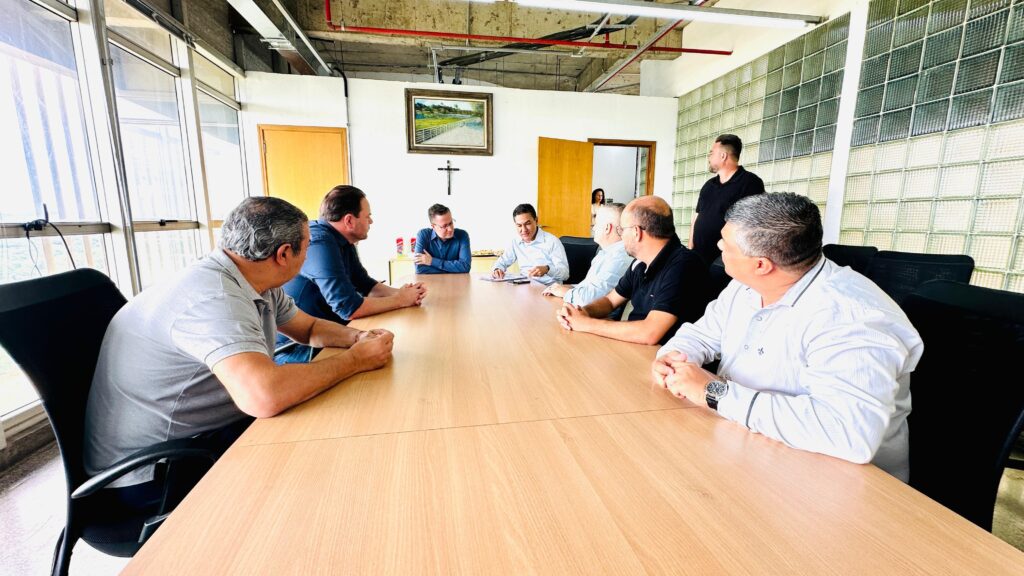 A imagem mostra um grupo de homens sentados em torno de uma mesa de madeira em uma sala de reuniões. Há um quadro com uma pintura na parede, uma porta e uma janela grande com vista para a cidade. A sala parece ser um escritório, com iluminação artificial e alguns detalhes arquitetônicos interessantes, como as paredes de vidro quadriculado. Os homens estão vestidos de forma casual a semi-formal, com camisas de manga curta e longa. Alguns estão olhando uns para os outros, enquanto outros parecem estar concentrados em documentos ou conversando. A atmosfera geral sugere uma reunião de trabalho ou uma discussão informal entre colegas.
