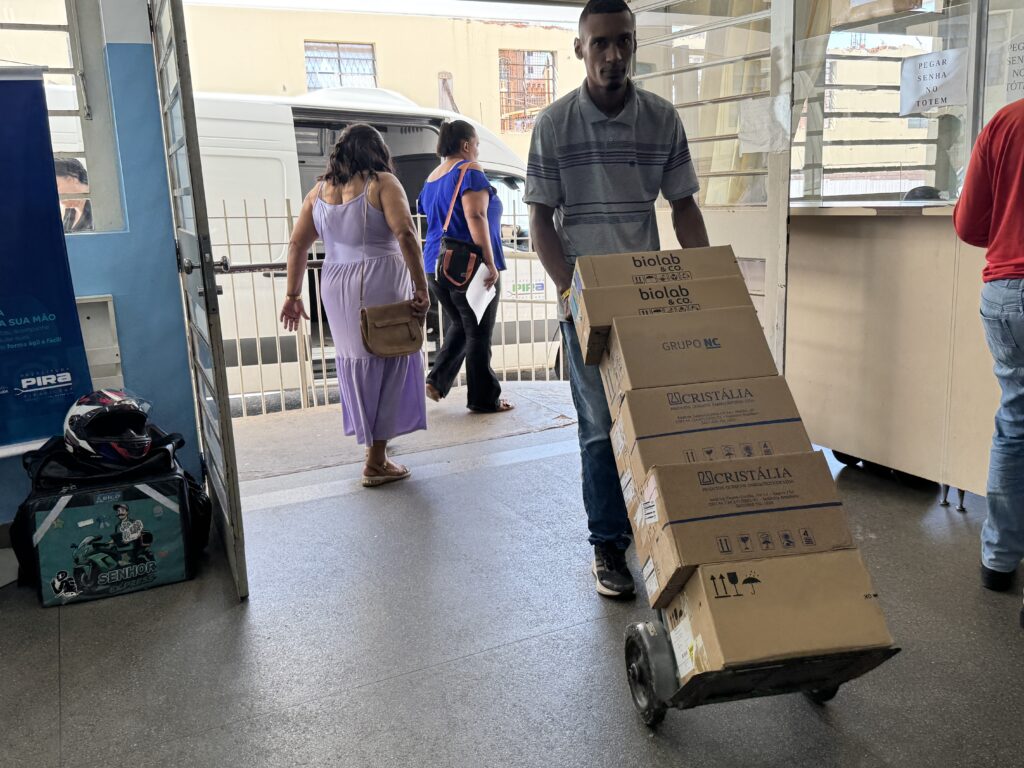A imagem mostra um homem empurrando um carrinho com várias caixas empilhadas. As caixas parecem ser de um laboratório ou empresa farmacêutica, com logotipos como "biolab & CO." e "CRISTÁLIA". Ele está em um ambiente interno, possivelmente uma recepção ou corredor, com outras pessoas ao fundo. À esquerda, há uma porta aberta com um cartaz azul preso à parede, que parece ser um anúncio de serviço. Ao lado do cartaz, há uma bolsa preta com um capacete de moto em cima, e uma bolsa menor com um adesivo de um entregador. No fundo, duas mulheres caminham para longe do homem. Uma delas usa um vestido lilás e a outra uma blusa azul. Há também uma van branca visível através de uma grade. No lado direito da imagem, há um balcão com um cartaz escrito "PEGAR SENHA NO TOTEM". Uma pessoa vestindo uma camisa vermelha está de pé perto do balcão, de costas para a câmera. O chão é de granito polido e as paredes são de cor clara. A iluminação sugere que seja dia.