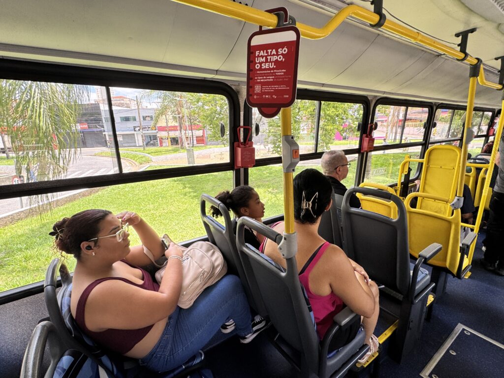 A imagem mostra o interior de um ônibus, com vários passageiros sentados. A iluminação é clara, vinda das janelas e do teto. No primeiro plano, à esquerda, uma mulher com óculos e fones de ouvido está sentada, segurando um celular e uma bolsa bege no colo. Ela veste uma blusa vinho e calça jeans. Ao lado dela, uma menina com cabelo preso está sentada, olhando para a frente. Mais atrás, um homem mais velho com óculos escuros está sentado. No centro e à direita, há assentos amarelos, típicos de ônibus, com algumas partes em plástico cinza. Uma placa vermelha com letras brancas está pendurada no teto, com a mensagem "FALTA SÓ UM TIPO, O SEU." e informações sobre doação de sangue. As janelas do ônibus mostram a vista do lado de fora, com árvores verdes, gramado e edifícios ao fundo. O ambiente parece ser urbano.