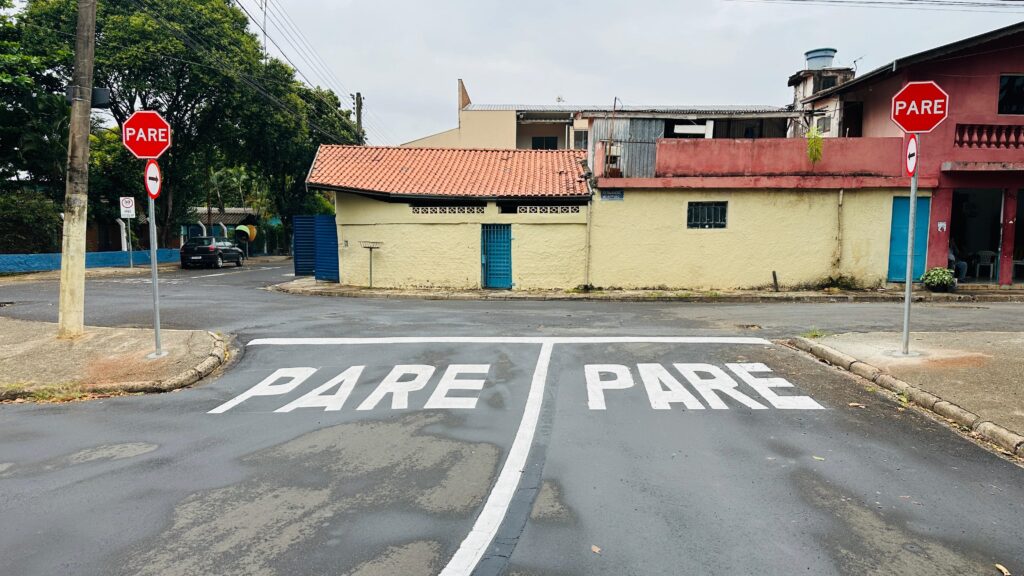 A imagem mostra uma intersecção de ruas com sinalização de "PARE" pintada no asfalto em ambas as direções. Há também placas de "PARE" hexagonais vermelhas em postes em ambos os lados da imagem. Ao fundo, vemos edifícios de cores variadas, com telhados de barro e algumas árvores. O céu está nublado, indicando um dia com pouca luz solar. Uma faixa branca divide a rua no primeiro plano, onde as palavras "PARE" estão escritas em letras brancas grandes.