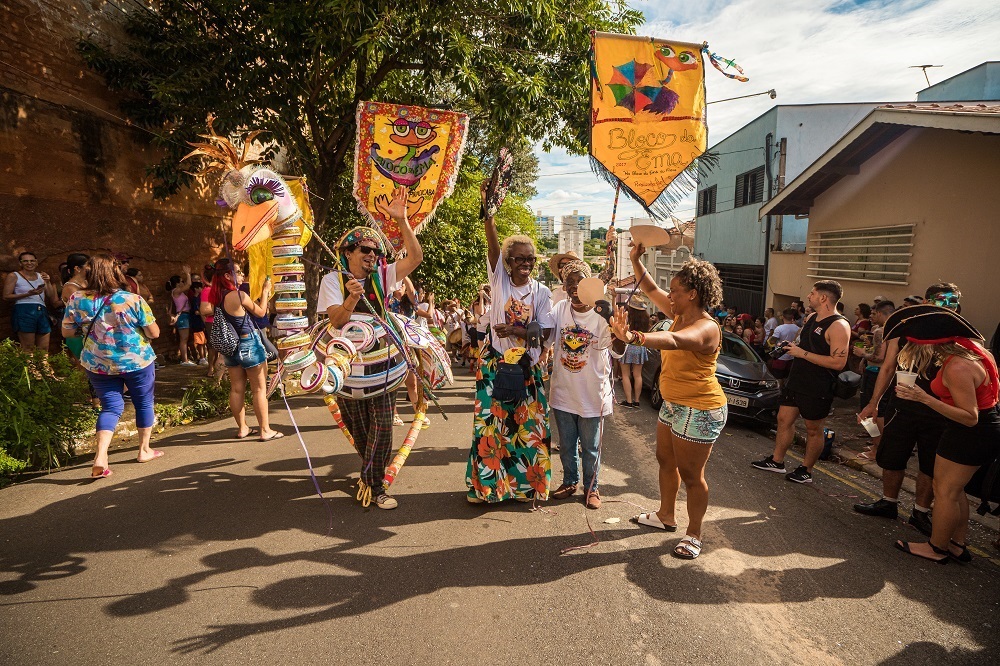 A imagem retrata um vibrante desfile de carnaval de rua, com pessoas fantasiadas e animadas. No centro da cena, um homem com uma fantasia elaborada, que lembra um pássaro com asas circulares e coloridas, acena para a câmera. Ele usa um chapéu adornado e óculos de sol. Ao seu lado, outras duas mulheres também participam da celebração, uma com um vestido longo estampado e a outra com uma camiseta branca, ambas sorrindo e acenando. Atrás deles, uma grande bandeira amarela com a inscrição "Bloco da Ema" e desenhos de guarda-chuvas coloridos é erguida. Mais adiante, outras pessoas com fantasias e adereços carnavalescos, algumas com instrumentos musicais, compõem o cenário festivo. A rua está ladeada por casas e árvores, e o céu azul com algumas nuvens indica um dia ensolarado. Pessoas assistem ao desfile pelas calçadas, criando uma atmosfera de comunidade e alegria.
