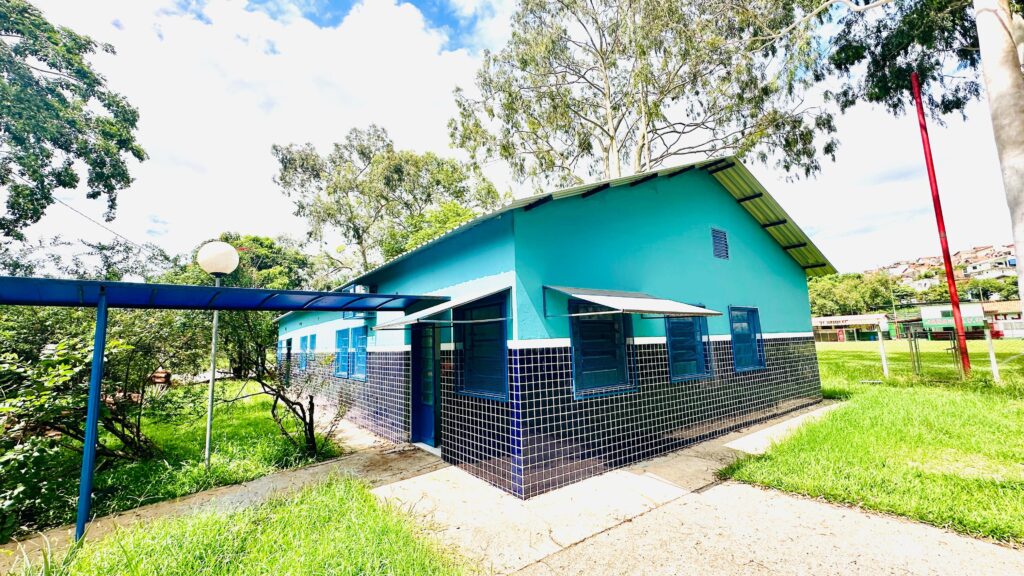 A imagem mostra um edifício de cor azul-turquesa, com detalhes em azulejos pretos e brancos na parte inferior. O edifício tem um telhado de metal inclinado e janelas azuis com persianas. Há uma estrutura azul coberta na frente do edifício, com um poste de iluminação esférico. O edifício está rodeado por relva verde e árvores frondosas. Ao fundo, avista-se um campo desportivo com graduação e algumas casas numa colina. O céu está parcialmente nublado com um sol radiante.