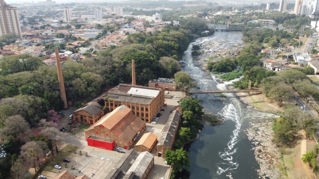 A imagem aérea mostra uma paisagem urbana com um rio sinuoso atravessando a cena. À esquerda, um complexo industrial com edifícios de tijolos vermelhos e uma alta chaminé de tijolos se destaca entre a vegetação exuberante. Várias chaminés industriais de tijolos se erguem no meio de árvores densas. À direita do rio, a paisagem urbana se estende, com casas residenciais e edifícios modernos ao fundo. Pontes conectam as margens do rio, e a água corre turbulenta sobre pedras, criando espuma branca. Carros e ruas podem ser vistos ao longo da margem direita. A atmosfera geral sugere uma área onde o desenvolvimento industrial convive com a natureza e a vida urbana.