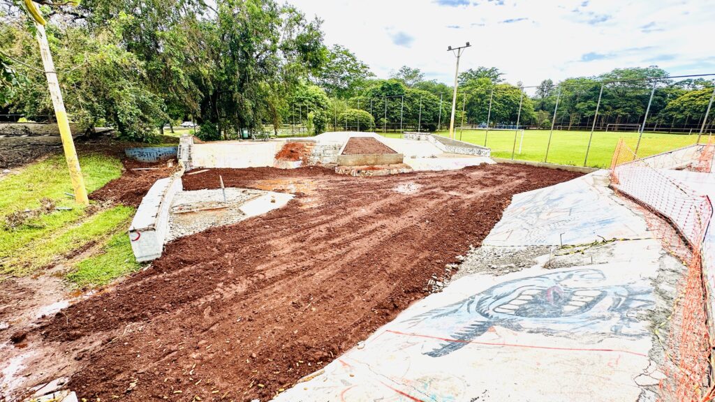A imagem mostra um skate park em processo de reforma. O chão está coberto de terra vermelha recém-cavada, com marcas de pneus visíveis, indicando que o material foi movimentado recentemente. Algumas estruturas de concreto do skate park, como rampas e bordas, ainda estão visíveis, mas parecem ter sido danificadas ou estão em processo de demolição/reconstrução. Ao redor da área principal do skate park, há grama verde e árvores frondosas, sugerindo que o local está situado em um parque ou área verde. Uma rede de proteção laranja delimita parte da área em construção. Ao fundo, é possível ver um campo gramado, possivelmente um campo de futebol, cercado por mais árvores e postes de iluminação. O céu está nublado, mas com alguns raios de sol.