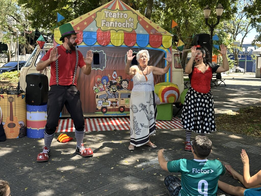 A imagem mostra um grupo de artistas de teatro de fantoches se apresentando ao ar livre. Em primeiro plano, um homem vestindo um chapéu verde, suspensórios xadrez, uma camisa vermelha e sapatos de palhaço grandes e vermelhos com estrelas brancas está em pose, com as mãos levantadas. Ao seu lado, uma mulher mais velha, com cabelos grisalhos curtos, usa um vestido branco com detalhes em renda e uma saia longa estampada com desenhos de âncoras e vida marinha. Do outro lado, uma mulher com um coque, usando um top vermelho rendado e uma saia preta com bolinhas brancas, também levanta as mãos. Ao fundo, há uma pequena tenda colorida com a inscrição "Teatro de Fantoche" e um desenho de uma Kombi com personagens animados. A tenda tem uma entrada com cortinas vermelhas e brancas listradas. A cena se passa em um ambiente externo, possivelmente um parque, com árvores ao fundo e o chão coberto por pedras. Alguns espectadores, incluindo um jovem vestindo uma camiseta verde com o número 9 e a palavra "Fictor>", estão sentados no chão, assistindo à apresentação. Há também equipamentos de som e elementos decorativos como instrumentos musicais de papelão e um dado gigante colorido.