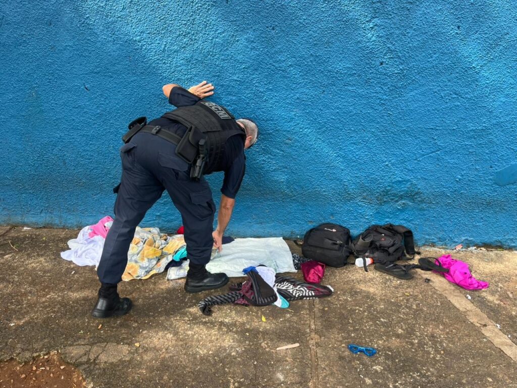 Um oficial de segurança, vestindo um uniforme azul escuro com um colete tático preto, está agachado em frente a uma parede azul vibrante. O colete tático tem a inscrição "GCM" em branco. O oficial está examinando um monte de roupas e pertences espalhados no chão de concreto. As roupas incluem peças de vestuário coloridas e brancas, algumas dobradas e outras desdobradas. Há também duas mochilas pretas, um par de chinelos pretos, uma bolsa rosa brilhante e um par de óculos de sol azuis no chão. O oficial parece estar em meio a uma investigação ou catalogação dos itens encontrados.
