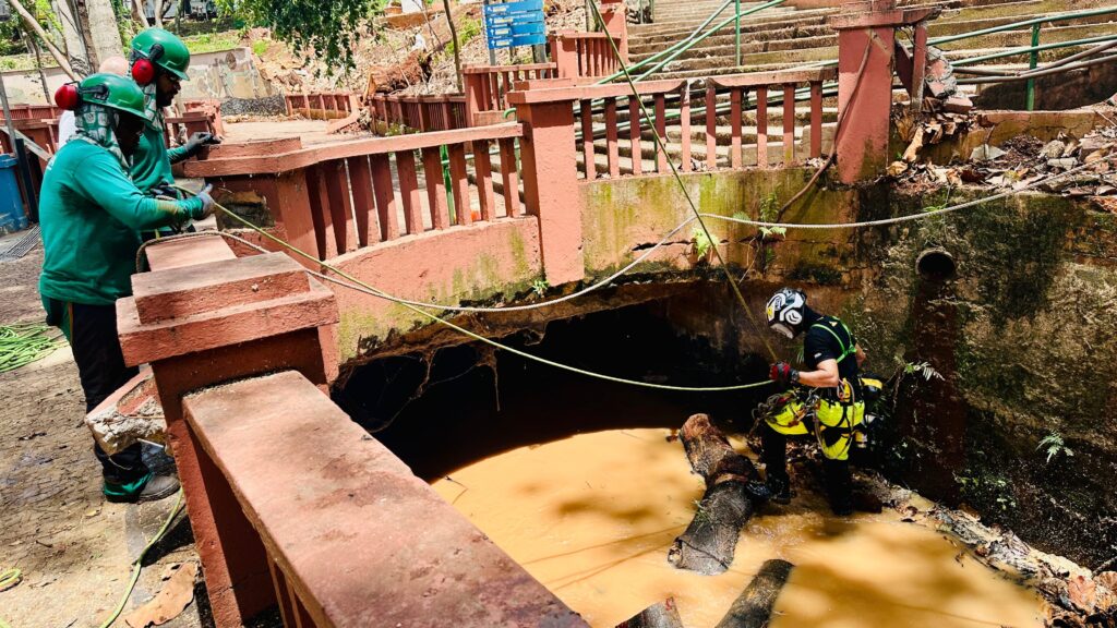 A imagem mostra uma cena de resposta a um desastre, possivelmente causada por um deslizamento de terra ou enchente. No centro, há uma grande quantidade de água barrenta, indicando alagamento ou lama acumulada. Três pessoas estão envolvidas na operação: duas, em primeiro plano, vestem camisas verdes, capacetes e protetores auriculares, e estão segurando cordas; a terceira, ao fundo, está dentro da água, usando equipamento de segurança completo, incluindo capacete e arnês, e parece estar realizando algum tipo de resgate ou limpeza. Ao fundo, vê-se um cenário de destruição, com uma estrutura de concreto danificada, possivelmente uma escada ou rampa, e bastante lama e entulho espalhados. A vegetação ao redor está parcialmente coberta pela lama. A cena transmite um esforço de recuperação em um ambiente difícil, com muita sujeira e riscos evidentes, e mostra a dedicação das equipes que trabalham para controlar os danos causados pelo desastre natural.