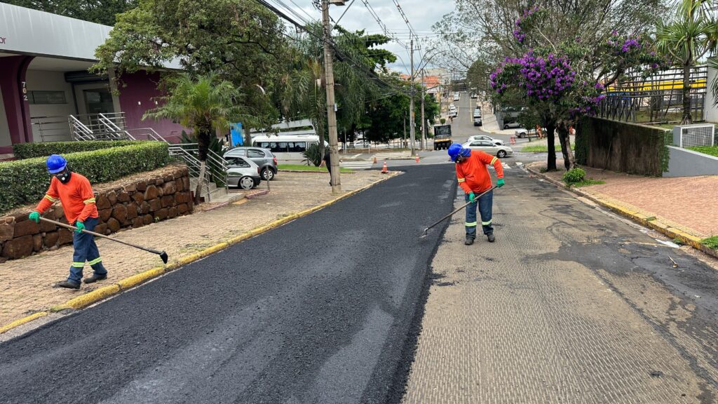 A imagem mostra dois trabalhadores em uma rua, realizando trabalhos de pavimentação asfáltica. Ambos estão vestidos com uniformes de segurança, incluindo capacetes azuis, luvas verdes e roupas de alta visibilidade em laranja. Um dos trabalhadores, mais próximo do espectador, está usando uma máscara e empunhando uma ferramenta longa com uma ponta larga, semelhante a um ancinho, para espalhar o asfalto recém-colocado. O outro trabalhador, mais distante, está fazendo um trabalho semelhante. O asfalto fresco cobre uma parte significativa da rua, com uma borda amarela demarcando o limite da área pavimentada. Ao fundo, a cena revela um ambiente urbano com edifícios, árvores (uma delas com flores roxas vibrantes), postes de eletricidade e tráfego de veículos. Há também um ônibus branco estacionado em um lote adjacente, e carros circulando na rua em declive. A calçada em primeiro plano é feita de pedras e a área ao lado da rua tem um muro de contenção de pedras. O céu está nublado, sugerindo um dia com condições climáticas amenas para o trabalho de pavimentação.