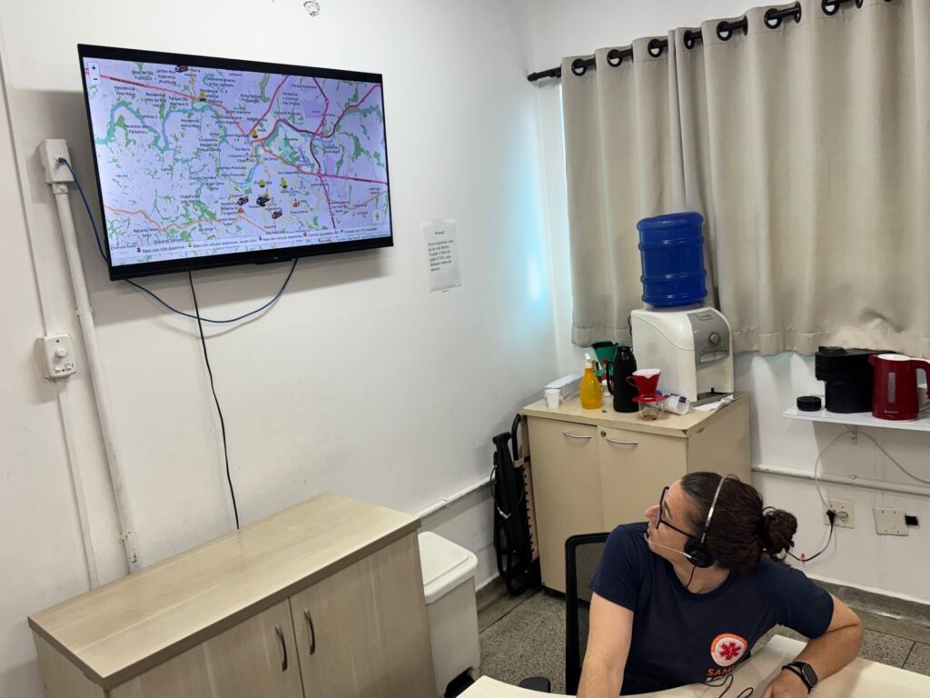 A imagem mostra um escritório com uma pessoa usando fones de ouvido, sentada em uma cadeira em frente a uma mesa. Há um monitor grande na parede exibindo um mapa com vários ícones, possivelmente indicando a localização de veículos ou unidades de serviço. Ao lado do monitor, há uma cortina e um bebedouro com um galão de água azul. Na mesa, há uma garrafa amarela, uma caneca preta, um filtro de café e outros itens. A pessoa parece estar trabalhando em um centro de operações ou de controle.