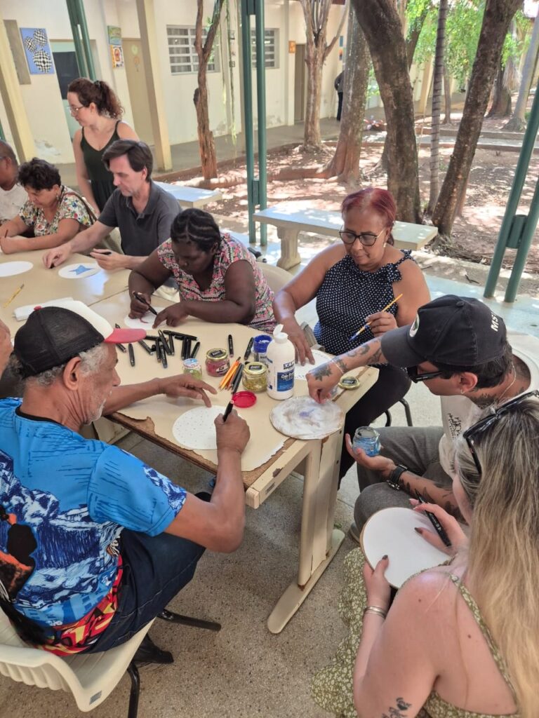 A imagem mostra um grupo de pessoas sentadas em volta de uma mesa, em um ambiente externo com árvores ao fundo. Todos parecem estar participando de uma atividade artística, possivelmente uma oficina de artesanato ou pintura. Na mesa, há materiais como potes de glitter, cola, pincéis e papéis. As pessoas estão concentradas em seus trabalhos, algumas desenhando ou pintando em papéis redondos, outras manuseando potes de glitter e cola. Há uma diversidade de idades e etnias no grupo. O ambiente sugere um local ao ar livre, talvez um pátio escolar ou um espaço comunitário, com mesas e bancos de madeira. A luz do sol ilumina a cena, criando algumas sombras. A atmosfera geral é de colaboração e criatividade.